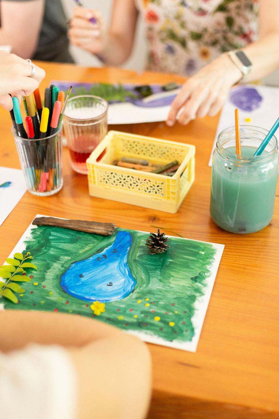 A painting of a blue pond in a green field, with a pine cone and leaves placed on it